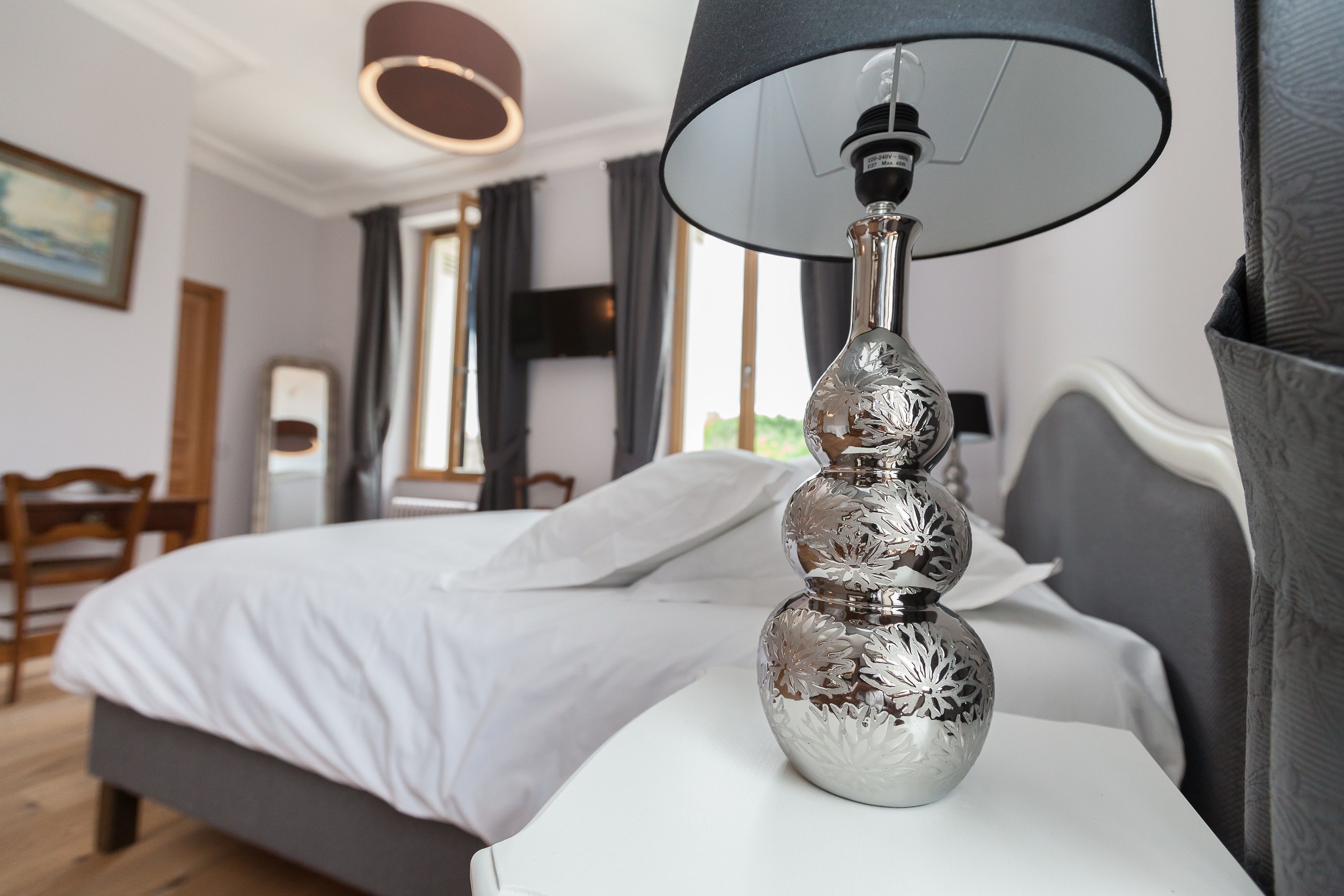 Ein ordentlich bezogenes Bett mit weißen Bettwäsche, eine silberne Vase mit schwarzem Lampenschirm auf einem Nachttisch und ein Holzstuhl im Hintergrund.