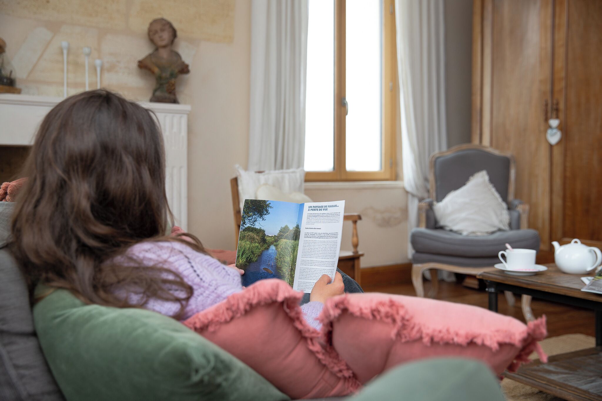 Una persona leyendo un libro en una habitación con chimenea, ventana y sillón.