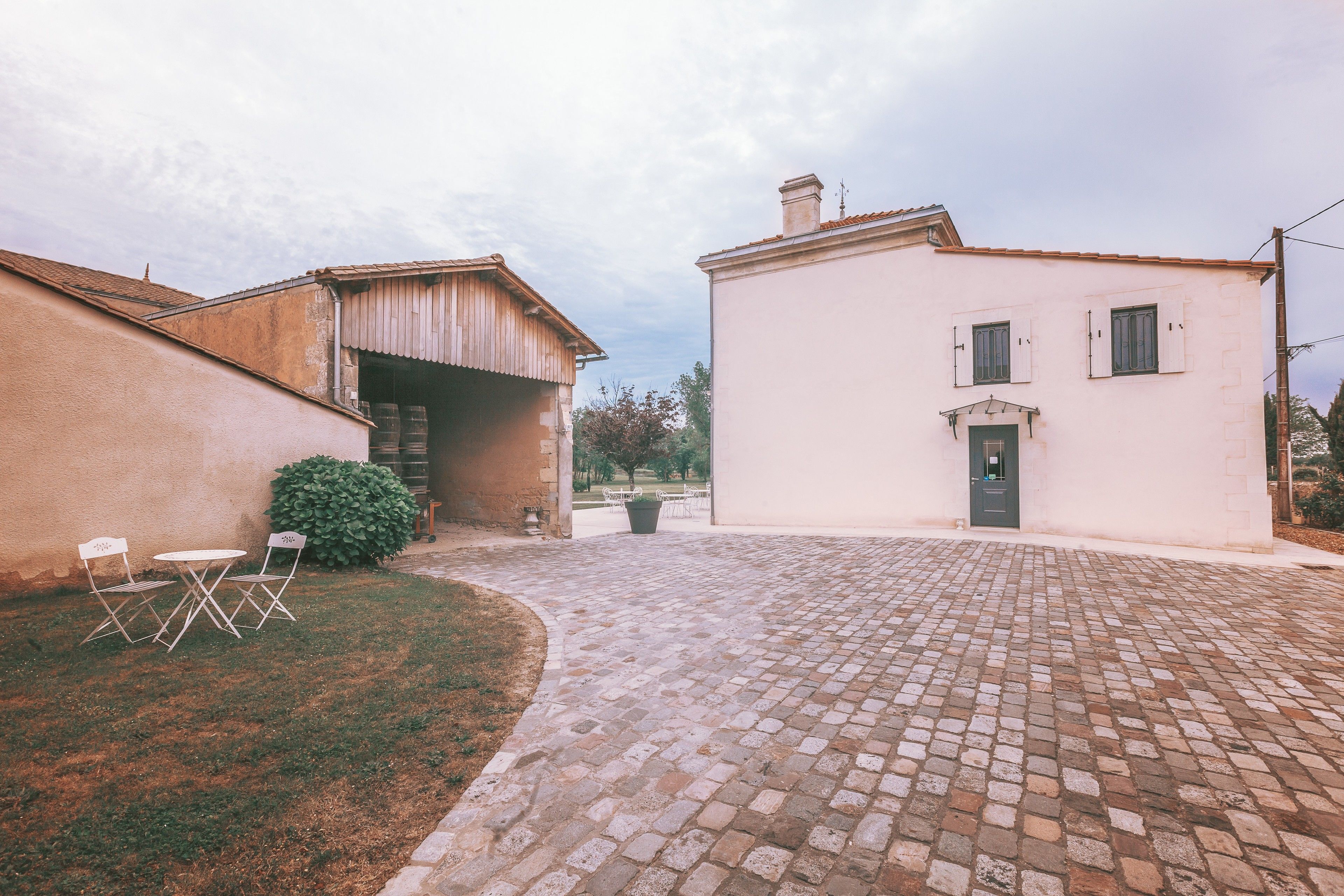 Un patio con área pavimentada, un edificio blanco con puerta y ventanas, y un pequeño jardín con mesa y sillas.