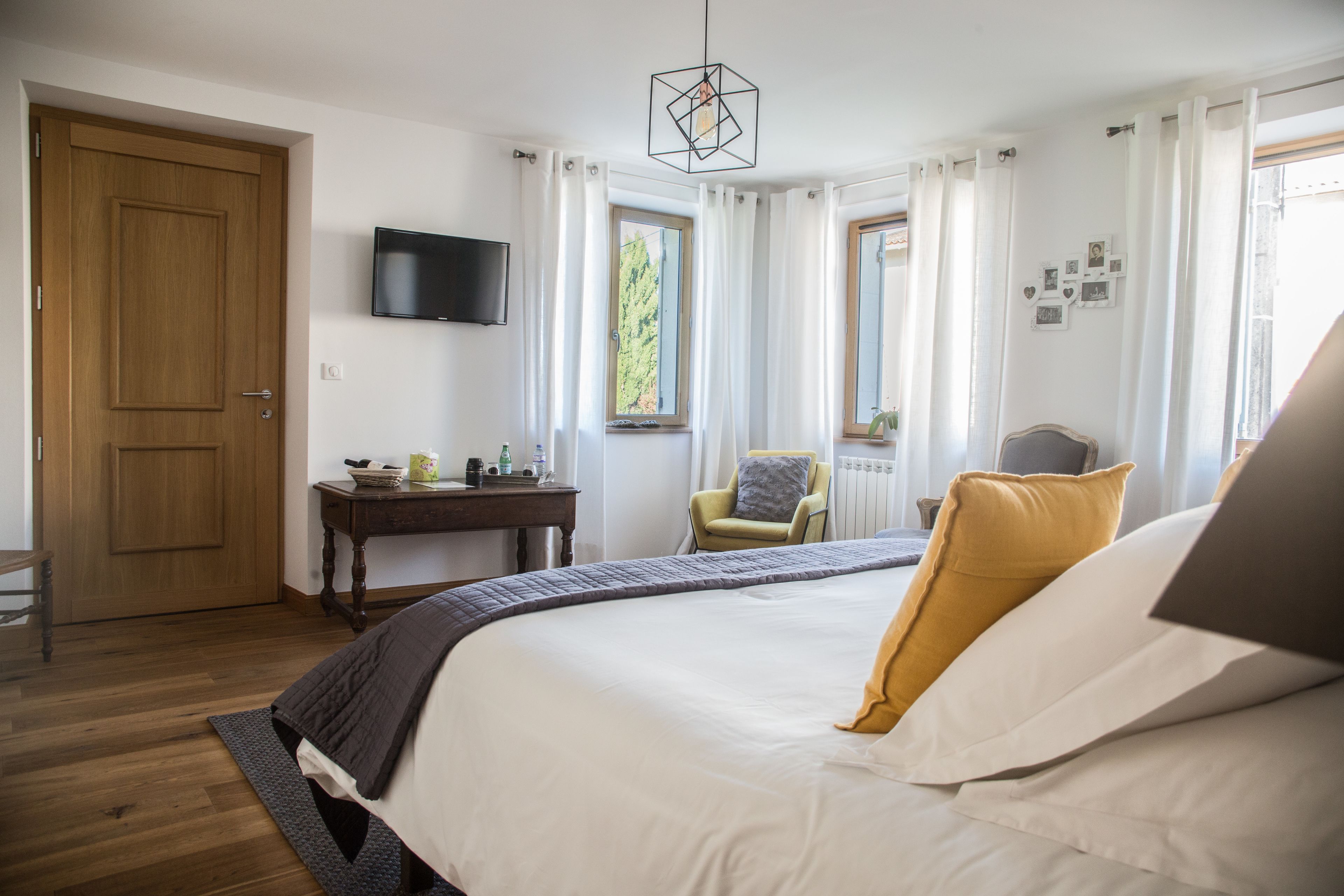 Un dormitorio con una cama grande, una puerta de madera, un televisor en la pared, una ventana con cortinas y una mesita con objetos encima.
