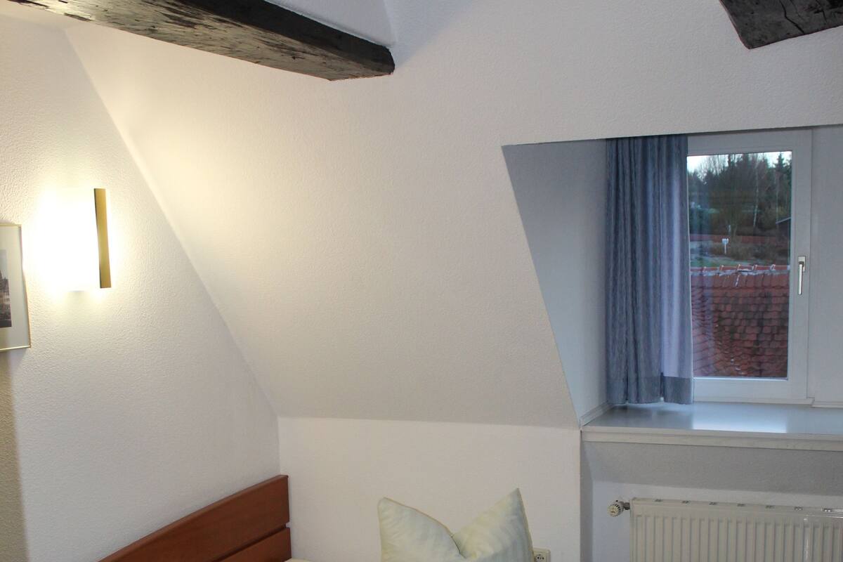 A small, neatly arranged bedroom with a single bed, a bedside table, a lamp, and a window with curtains.