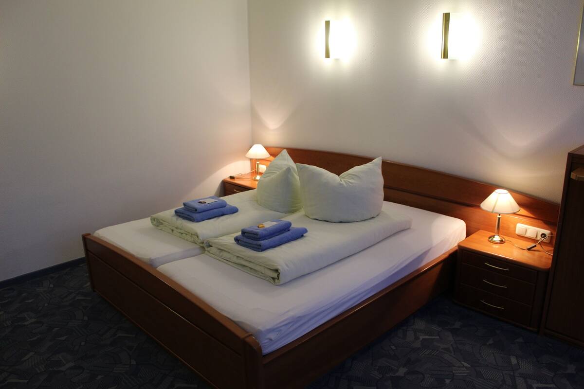 A neatly made bed with pillows and folded towels, two bedside lamps, and a wooden nightstand with drawers.