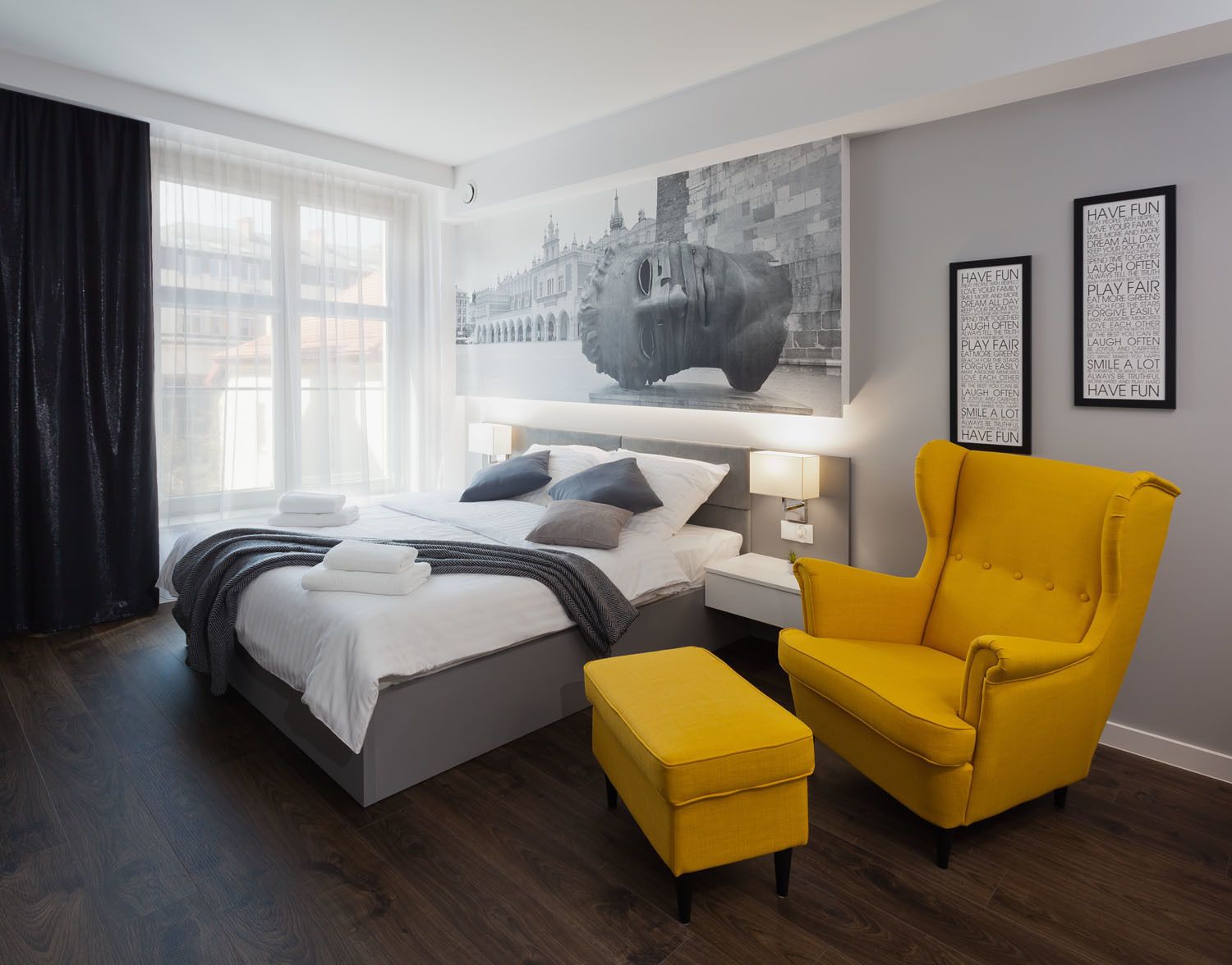 Quarto de hotel moderno com uma cama grande, uma poltrona amarela vibrante e um mural com vista para a cidade na parede.