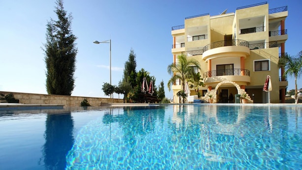A clear blue pool with a modern building featuring balconies and a stone wall in the background. - Apartment Emelia (Paphos)