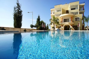 Una piscina azul cristalina con un edificio moderno que cuenta con balcones y un muro de piedra al fondo.