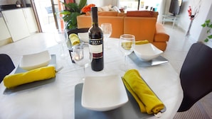 A dining table set with a bottle of wine, glasses, and plates, with a sofa and TV in the background. - Apartment Emelia (Paphos)
