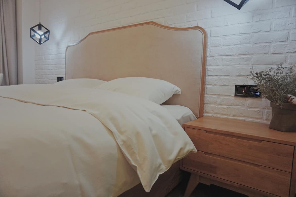 Un lit avec une tête de lit en bois, une table de chevet avec une plante et un mur en briques blanches.