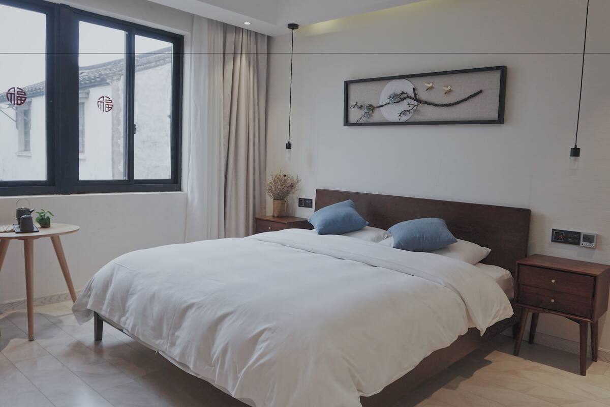 Une chambre à coucher moderne avec un grand lit, deux tables de chevet, une petite table avec une plante et une fenêtre avec des rideaux.
