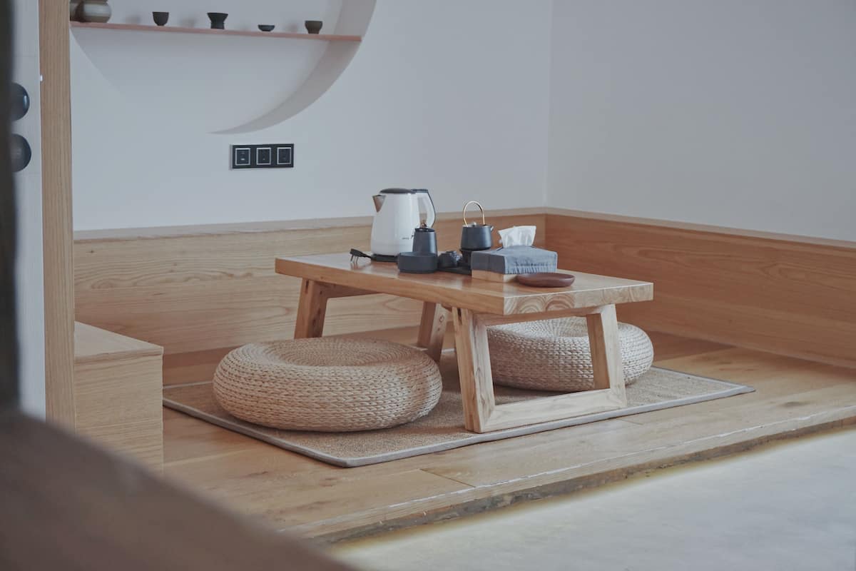 Une table en bois, sur laquelle sont posés une bouilloire, une théière et des tasses, est placée sur un tatami, avec deux poufs en osier à proximité.