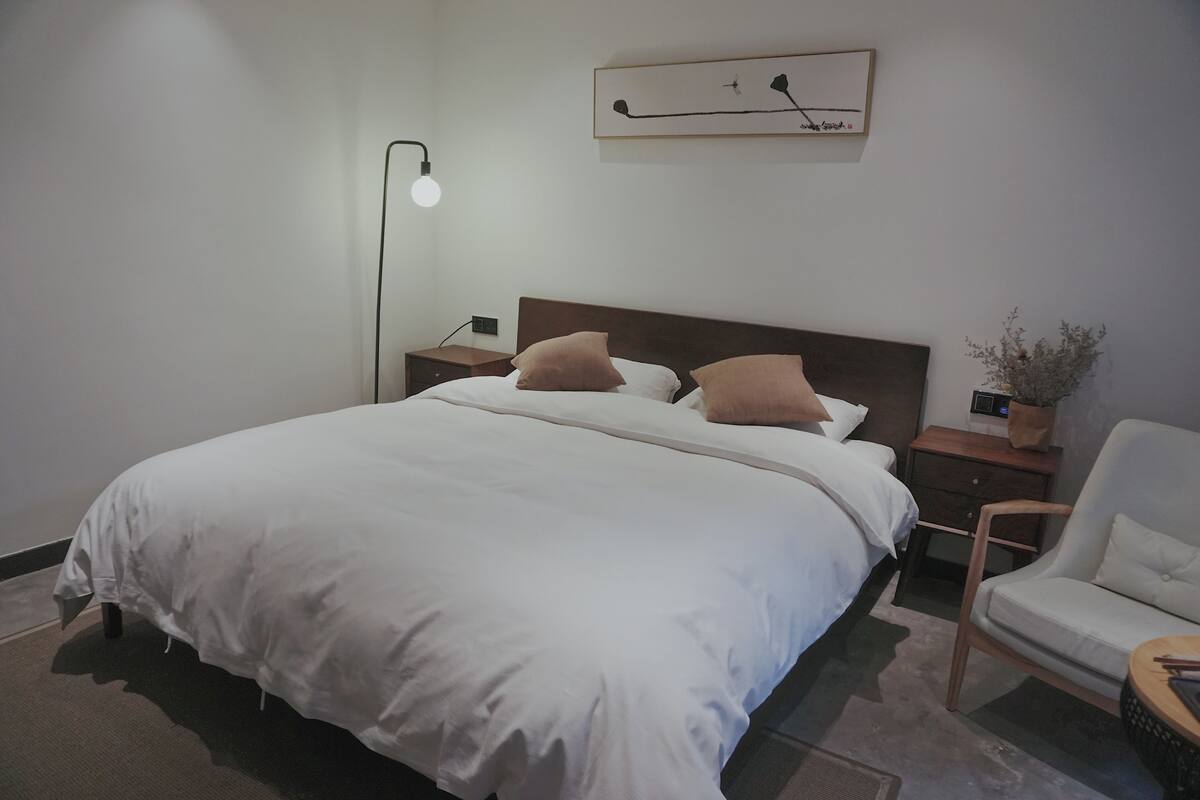 Une chambre d’hôtel bien aménagée, avec un grand lit, une table de chevet avec une lampe, une chaise et une petite table sur laquelle est posé un vase.