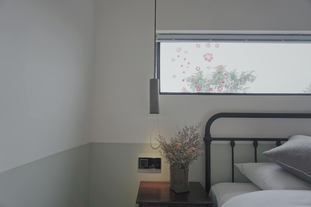 Une chambre à coucher avec un lit, une table de chevet ornée d’un vase de fleurs, une lampe fixée au mur et une fenêtre donnant sur des arbres et des fleurs.