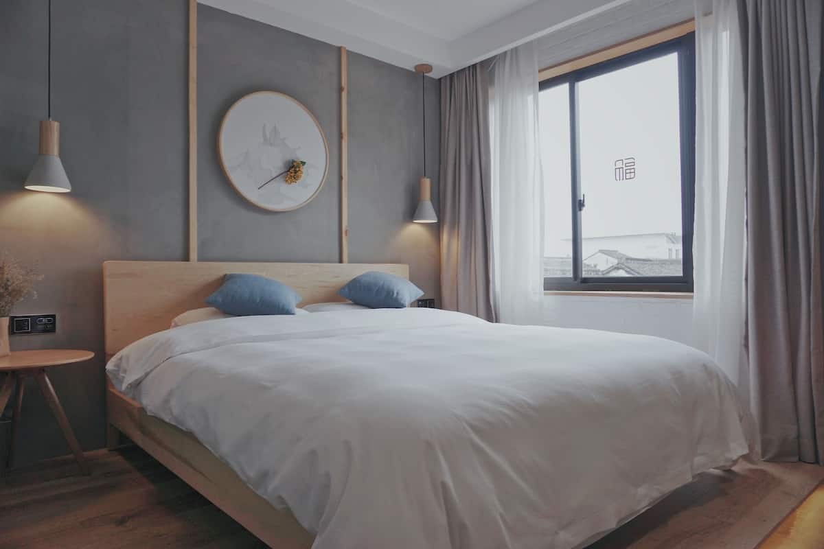 Une chambre d’hôtel moderne avec un grand lit, deux lampes de chevet, une horloge murale ronde et une fenêtre avec des rideaux.
