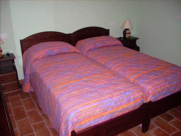 A double bed with a striped bedspread, a wooden headboard, a nightstand with a lamp, and a dresser with a mirror. - Agriturismo I Getsemani (Bevagna)