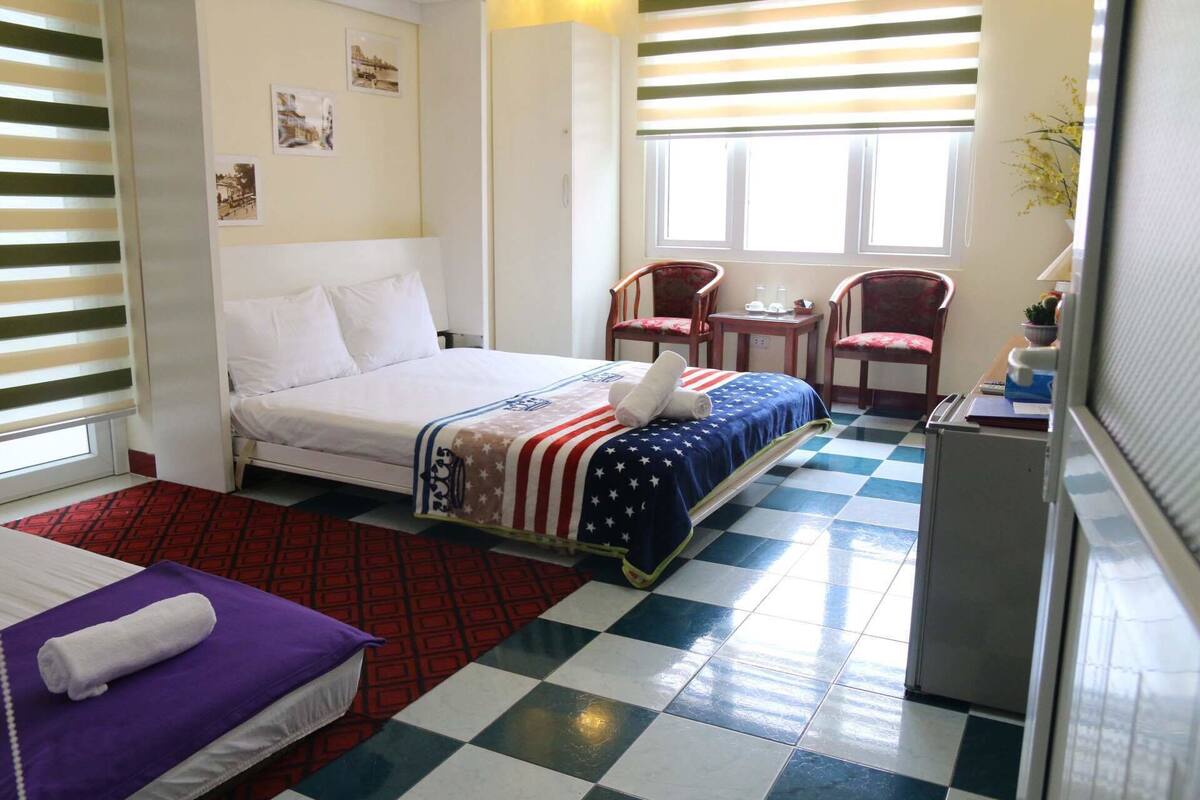 Habitació d'hotel amb un llit, un escriptori petit, dues cadires, una finestra amb persianes i un terra de pissos quadriculats.