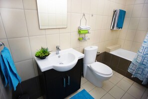 A bathroom with a white sink, a toilet, and a bathtub. There is a blue towel on the floor and a green plant on the sink. - Apartment Sinogi (Paphos)