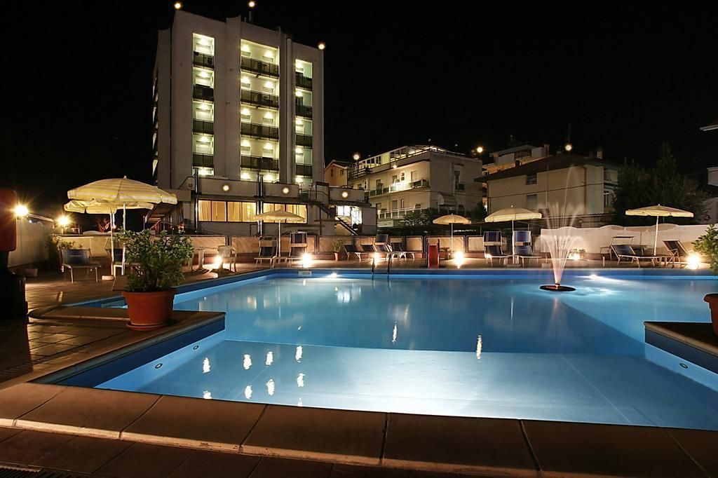 Una piscina con sillas de descanso y sombrillas, rodeada de edificios con ventanas iluminadas.