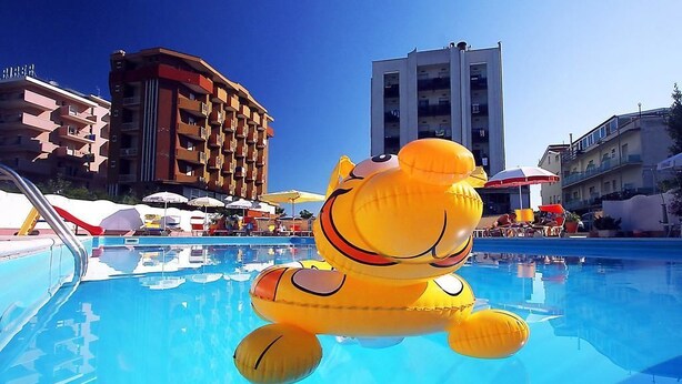 A pool area with a large inflatable animal float, surrounded by buildings and umbrellas. - Hotel Avila In (Rimini)