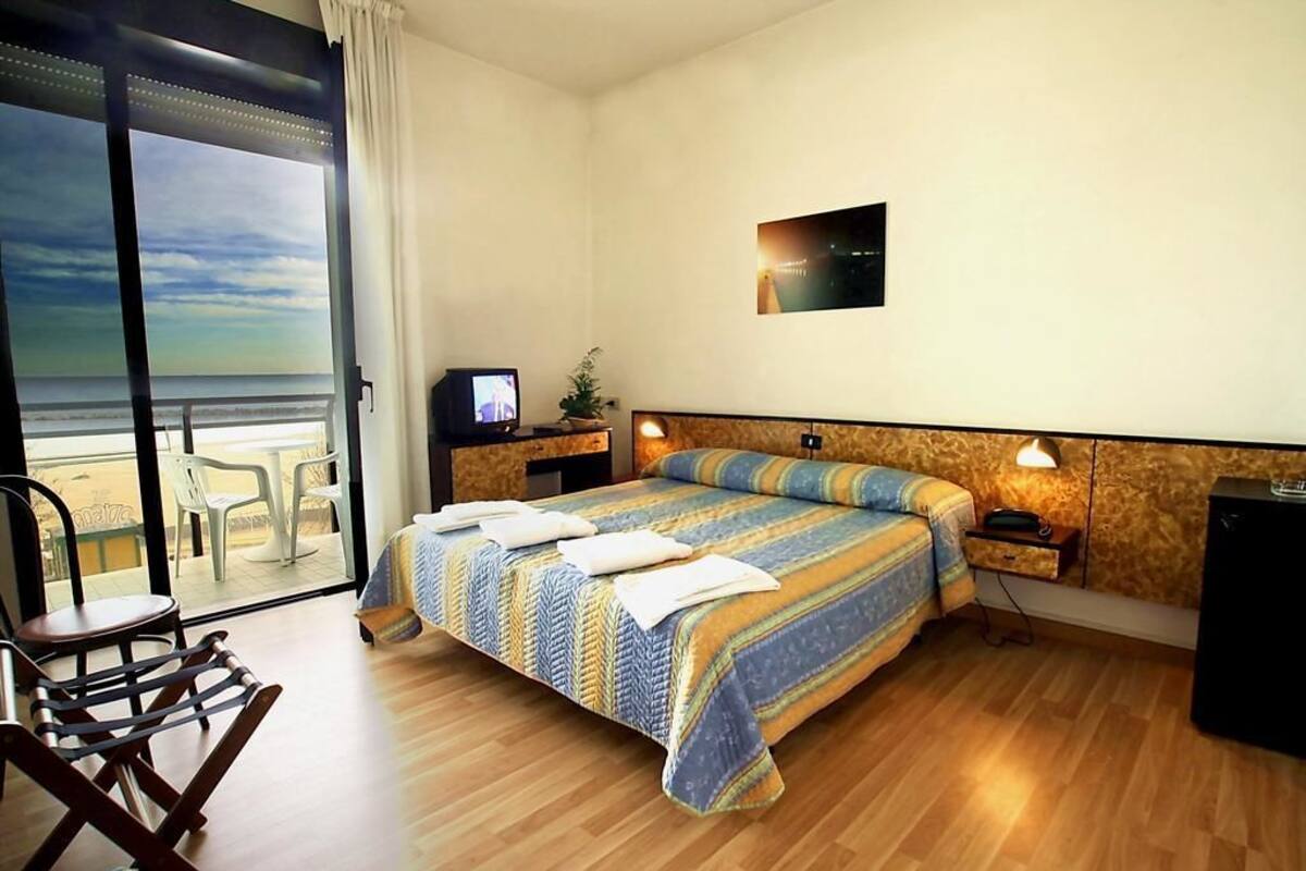 A hotel room with a large bed, a TV, a desk, and a view of the beach.