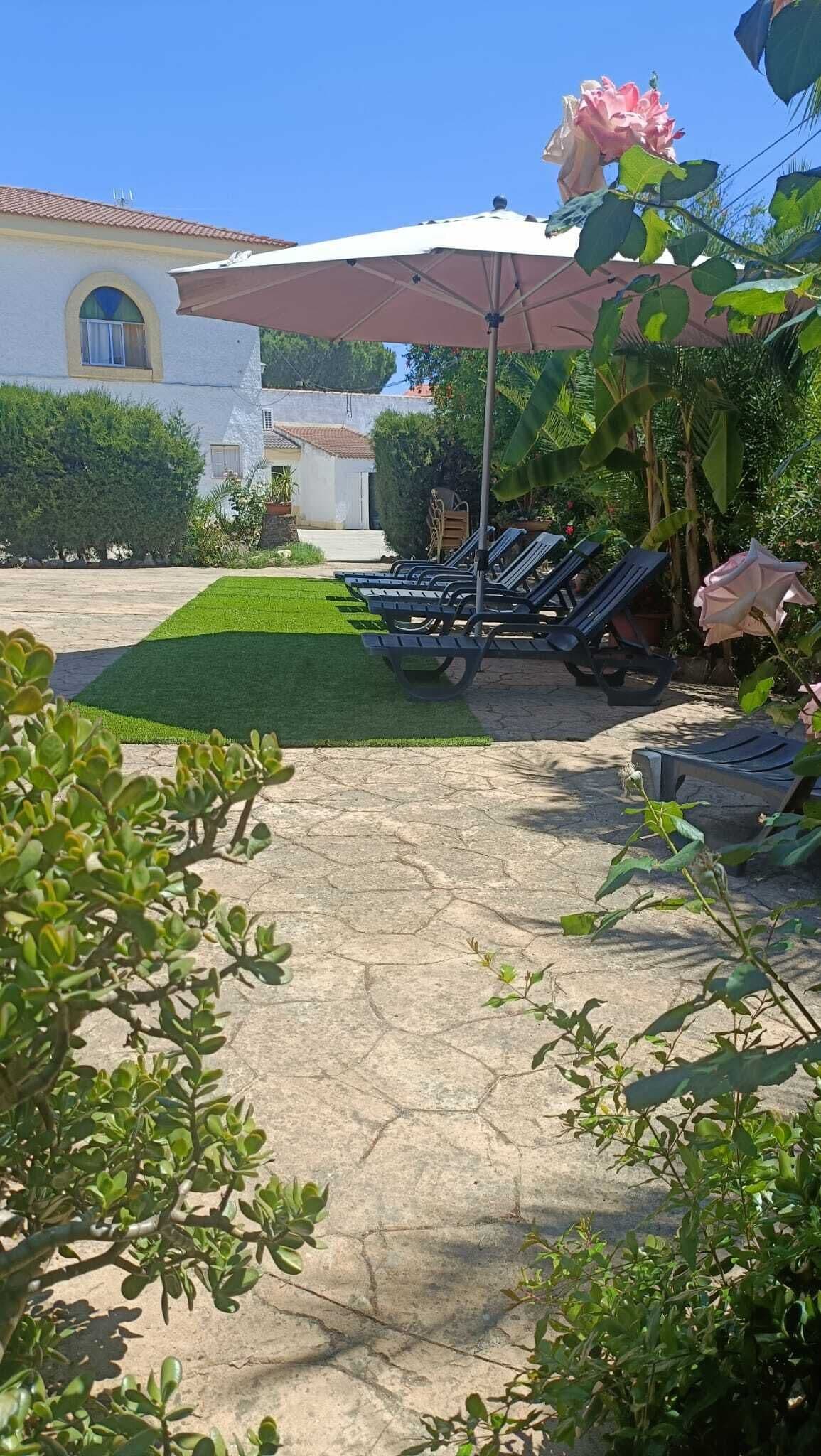 A patio area with lounge chairs and a large umbrella, surrounded by greenery and a building in the background.