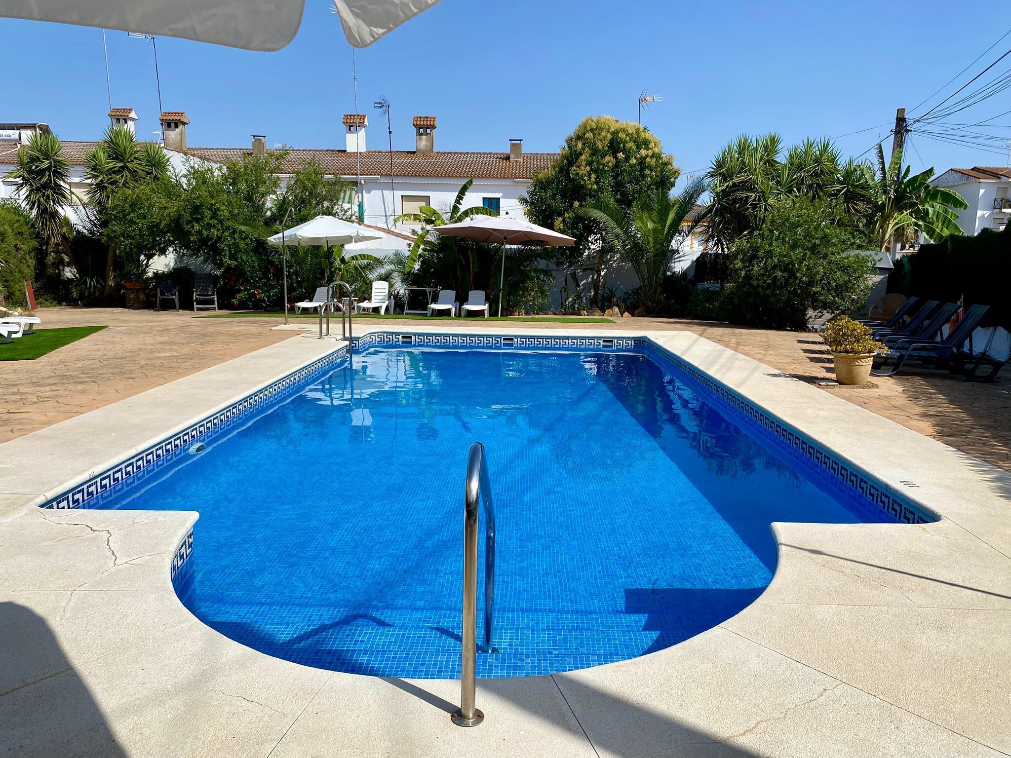 Una piscina con agua cristalina azul, rodeada por una terraza pavimentada, con sillas de descanso y sombrillas al fondo.