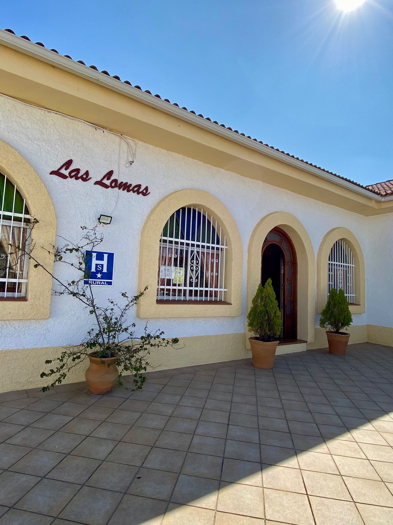 Un edificio con el letrero 'Las Lomitas' y un cartel azul que indica zona rural. Se observan tres ventanas de arco con rejas de metal, y dos macetas en primer plano.