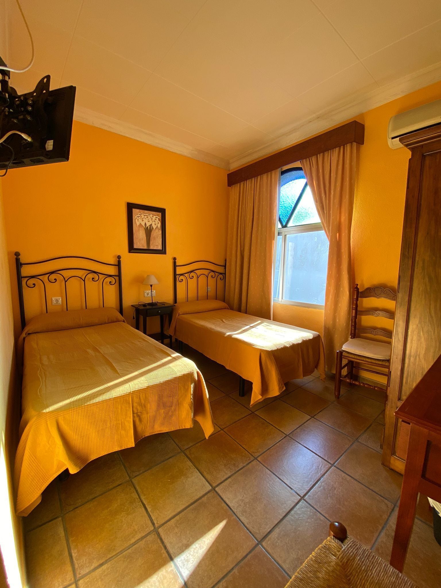 Une chambre avec deux lits, une chaise, un bureau, une fenêtre avec des rideaux et un tableau encadré au mur.