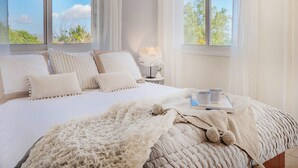 A bedroom with a large bed, a bedside table with a lamp, and a view of trees through the windows. - Villa Atlantica (Pegeia)