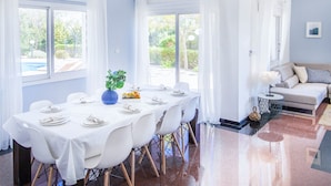 A dining area with a table set for six, large windows with curtains, a sofa, and a view of greenery outside. - Villa Atlantica (Pegeia)