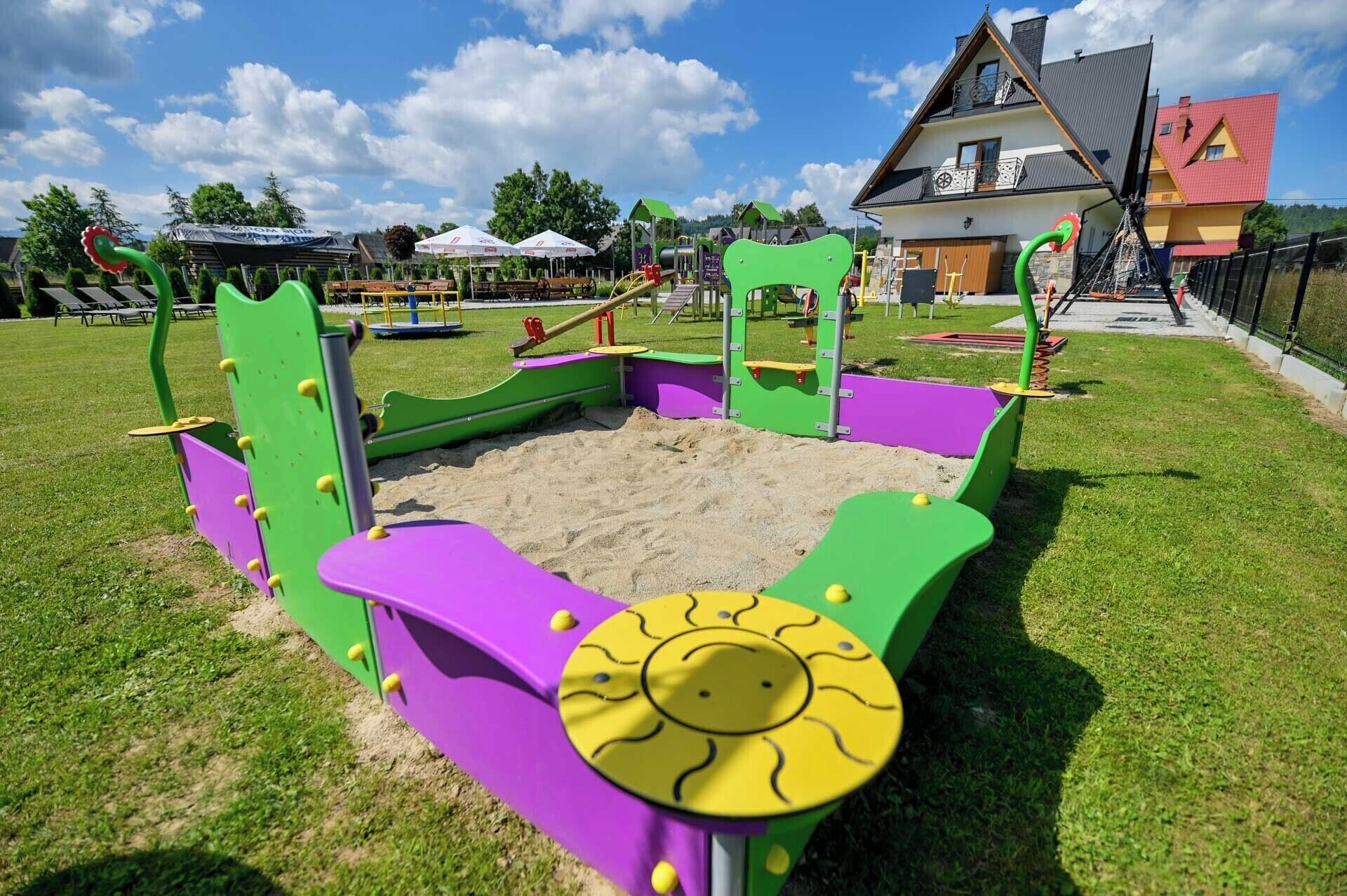 Un terrain de jeux avec un bac à sable et des structures d’escalade, entouré de pelouse et avec une maison en arrière-plan.