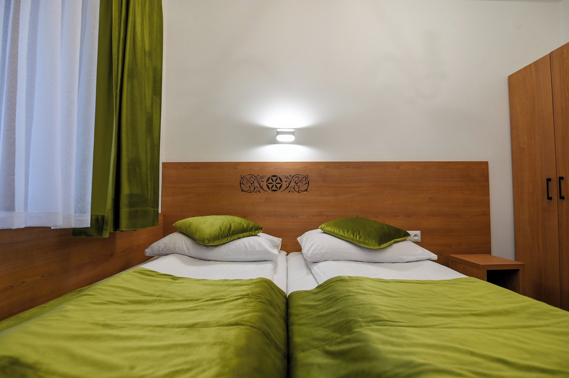 A double bed with green bedding, a wooden headboard with a decorative emblem, and a nightstand with a lamp.
