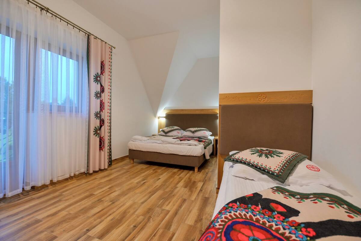 Un dormitorio con piso de madera, una cama con colcha colorida, cabecera con diseño decorativo y ventana con cortinas traslúcidas.