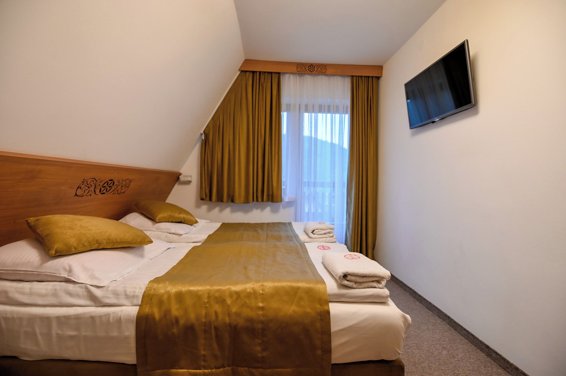 A hotel room with a bed, a headboard with a decorative design, a flat-screen TV mounted on the wall, and a window with curtains.