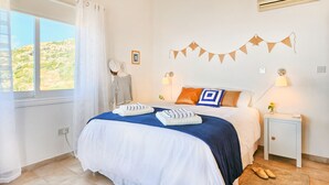 A neatly made bed with a blue and white bedspread, a white wall with a picture frame, a small bedside table with a lamp, and a window with curtains. - Villa Captains (Pegeia)