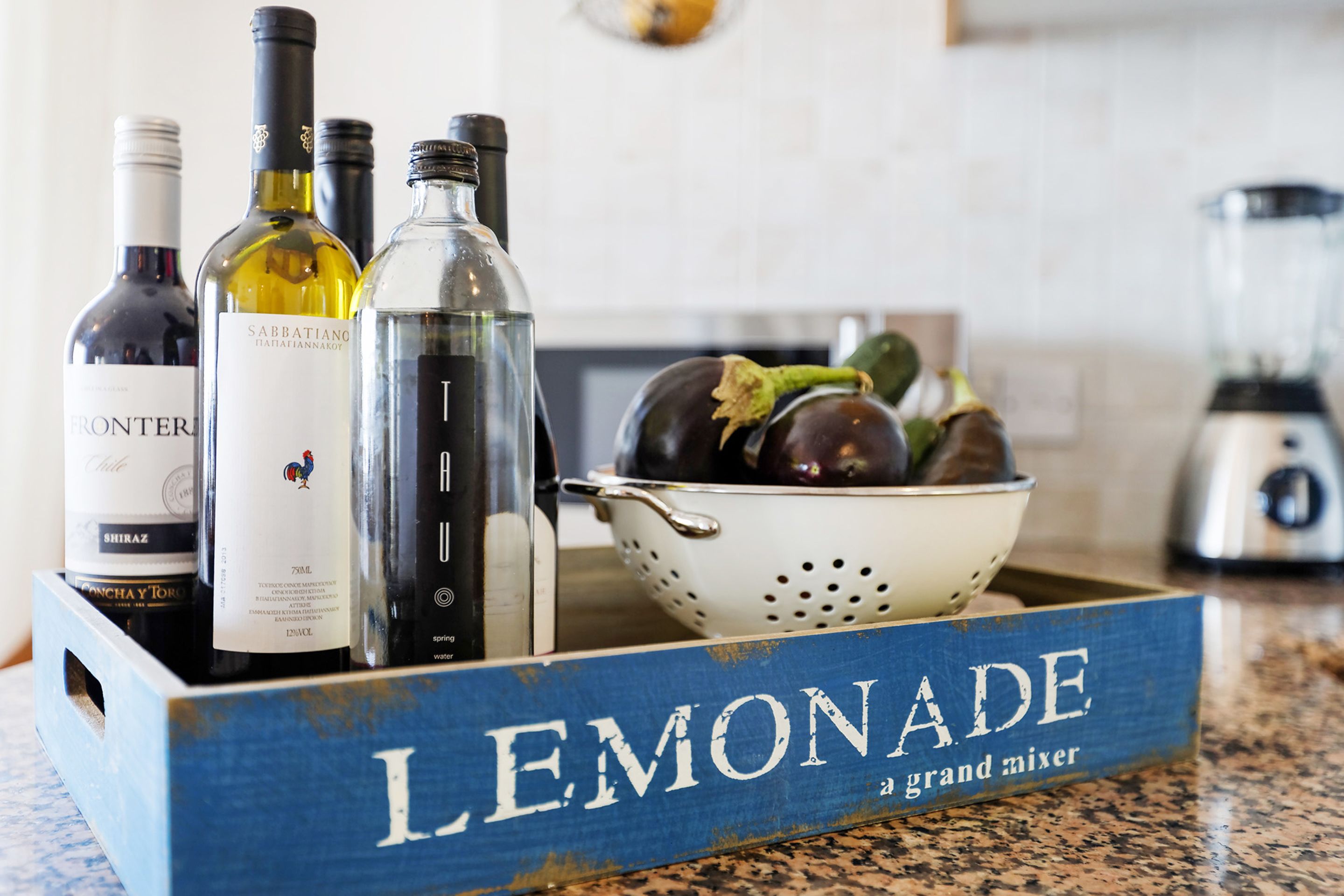 A wooden crate labeled "LEMONADE grand mixer" containing bottles of wine and a colander with vegetables.