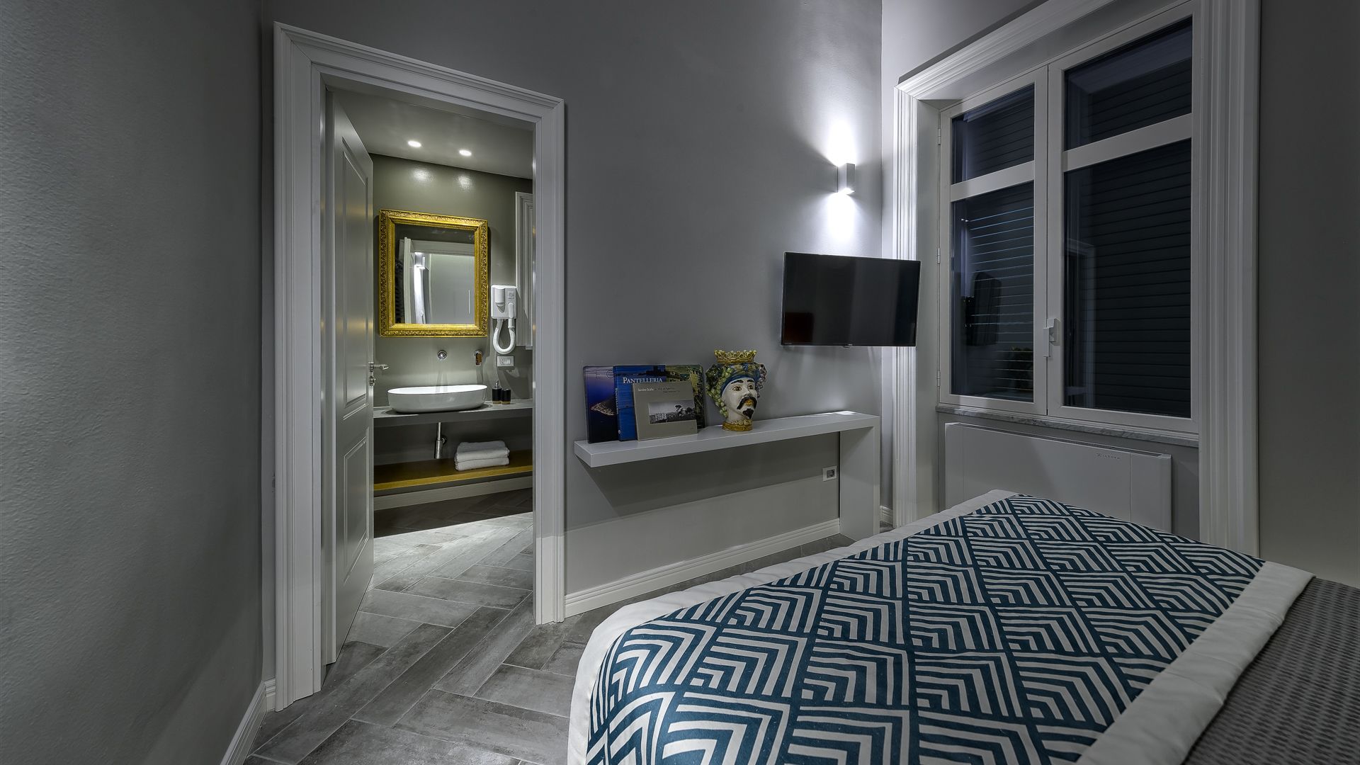 A bedroom with a bed, a nightstand, a shelf with books and a vase, a TV mounted on the wall, and a window with blinds.