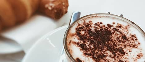 Uma chávena de café com uma espuma cremosa e um pouco de cacau em pó, acompanhada por um croissant sobre um prato branco.