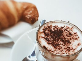 Una taza de café con espuma y un poco de cacao en polvo, junto a un croissant sobre un plato blanco.