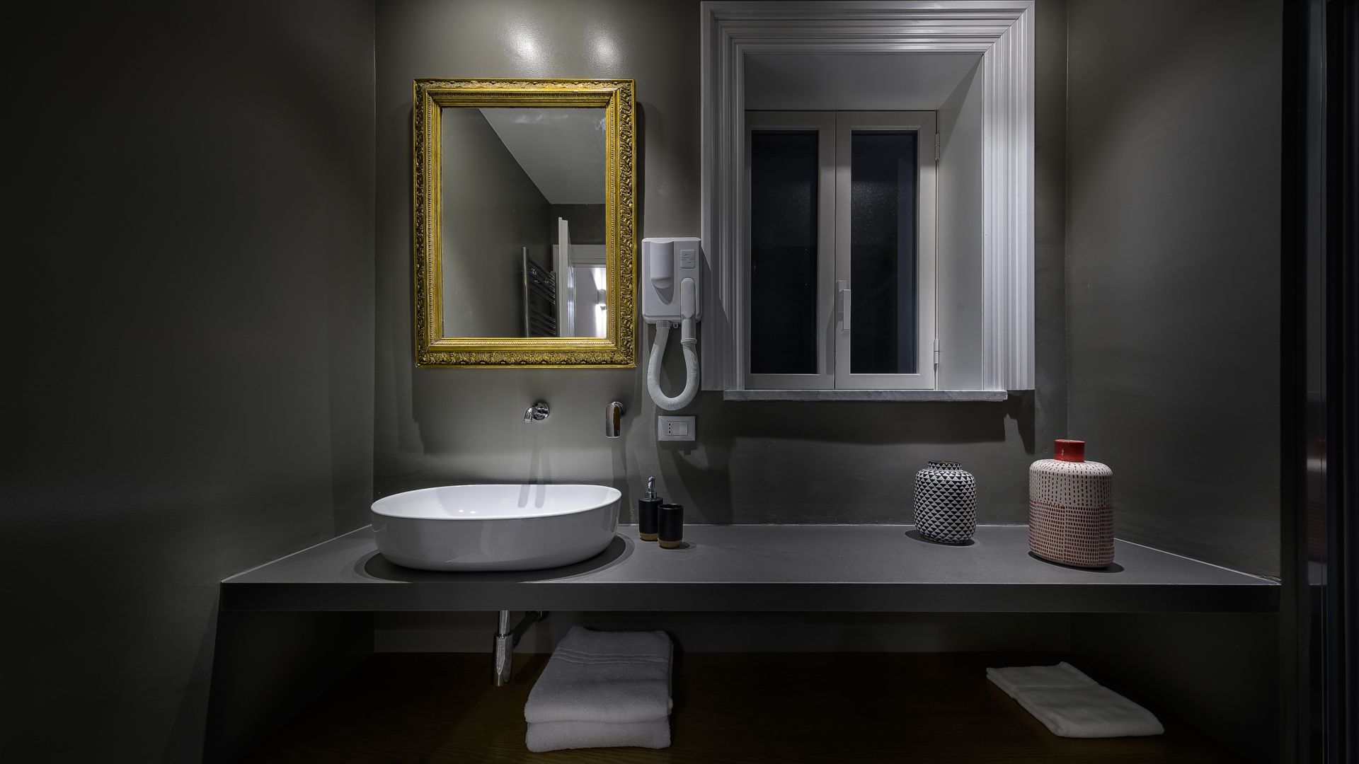 A modern bathroom with a white sink, a gold-framed mirror, a white toilet, and a window.