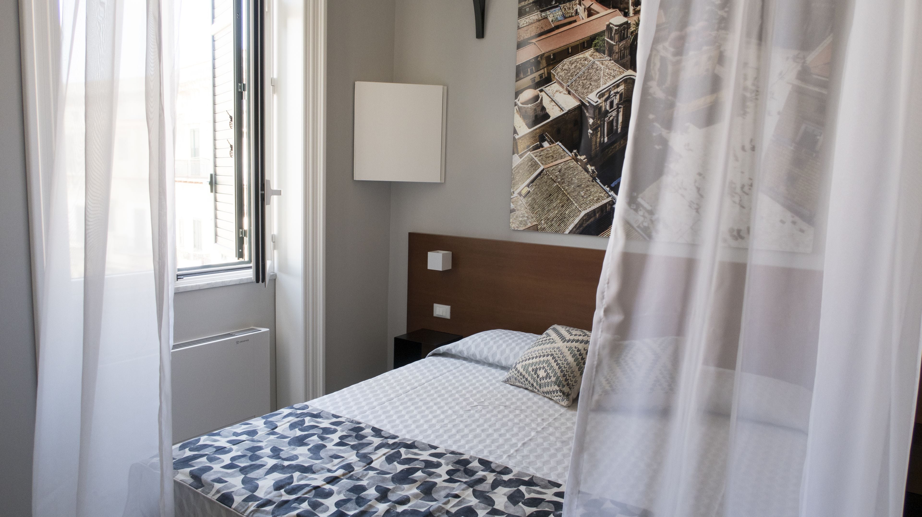 A hotel room with a bed, a window with sheer curtains, a wall-mounted light, and a large framed picture.