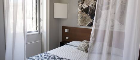 A hotel room with a bed, a window with sheer curtains, a wall-mounted light, and a large framed picture.