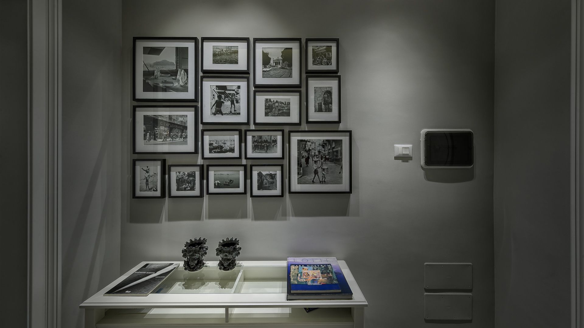 Una pared galería con fotografías blanco y negro enmarcadas, una vitrina de vidrio con un libro, y un termostato en la pared.