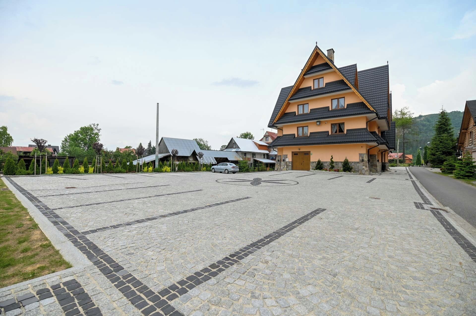 A building with a sloped roof, a parking area with a cobblestone pattern, and a few cars parked.