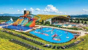 Parque acuático con múltiples toboganes y piscinas, un edificio grande con fachada de vidrio y una zona de césped con tumbonas.