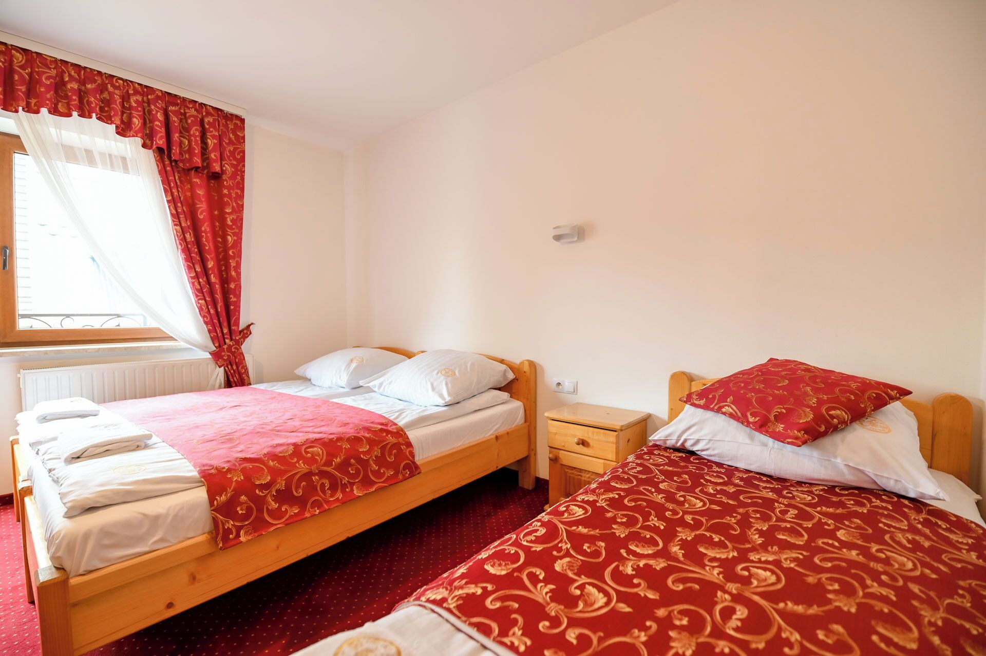 A bedroom with two beds, each with a red and white patterned bedspread. There is a window with red curtains, a bedside table with a lamp, and a radiator on the wall.