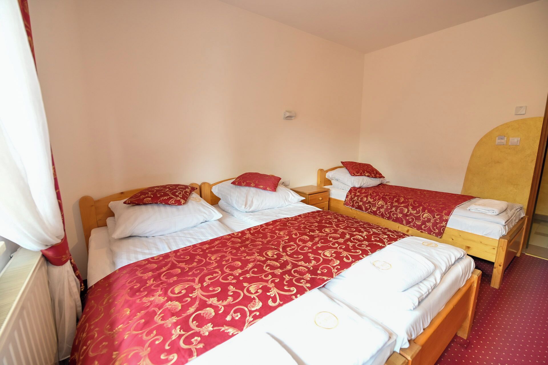 Ein Zimmer mit zwei Einzelbetten, beide mit Holz-Kopfteilen, weißer Bettwäsche und Decken mit rotem und goldenem Muster. An der Wand befindet sich ein Holzschrank und ein Heizkörper.