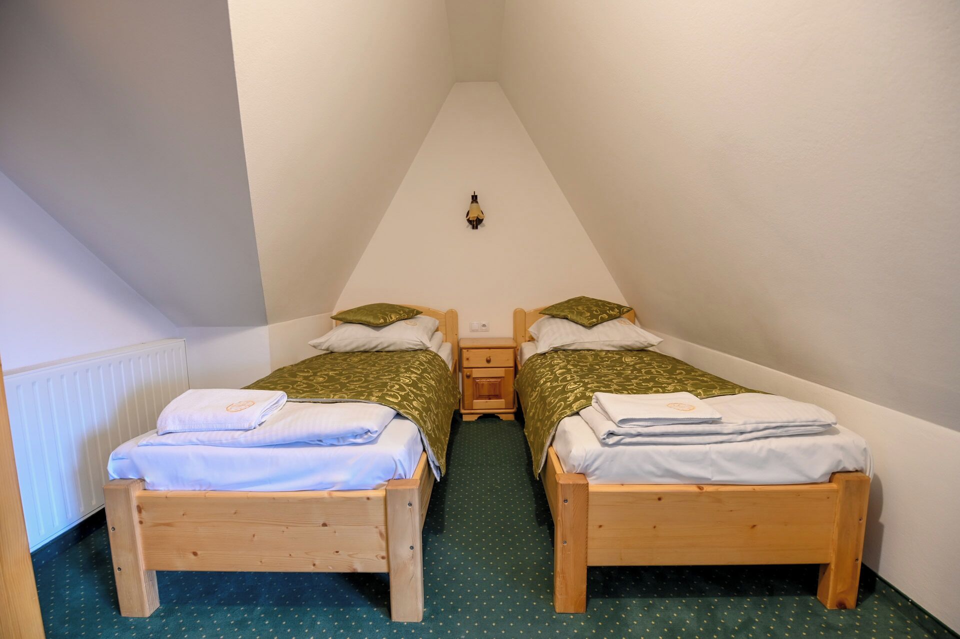 Una habitación pequeña y triangular con dos camas de madera, cada una con cubrecama verde y almohadas blancas. Hay una mesita de noche entre las camas, y una lámpara de pared sobre la mesita.