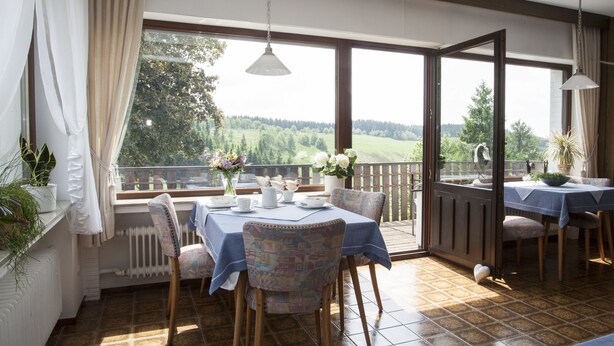 A dining area with a table set for two, a view of the outdoors, and a large window with curtains. - Pension Haus Brieden (Winterberg)