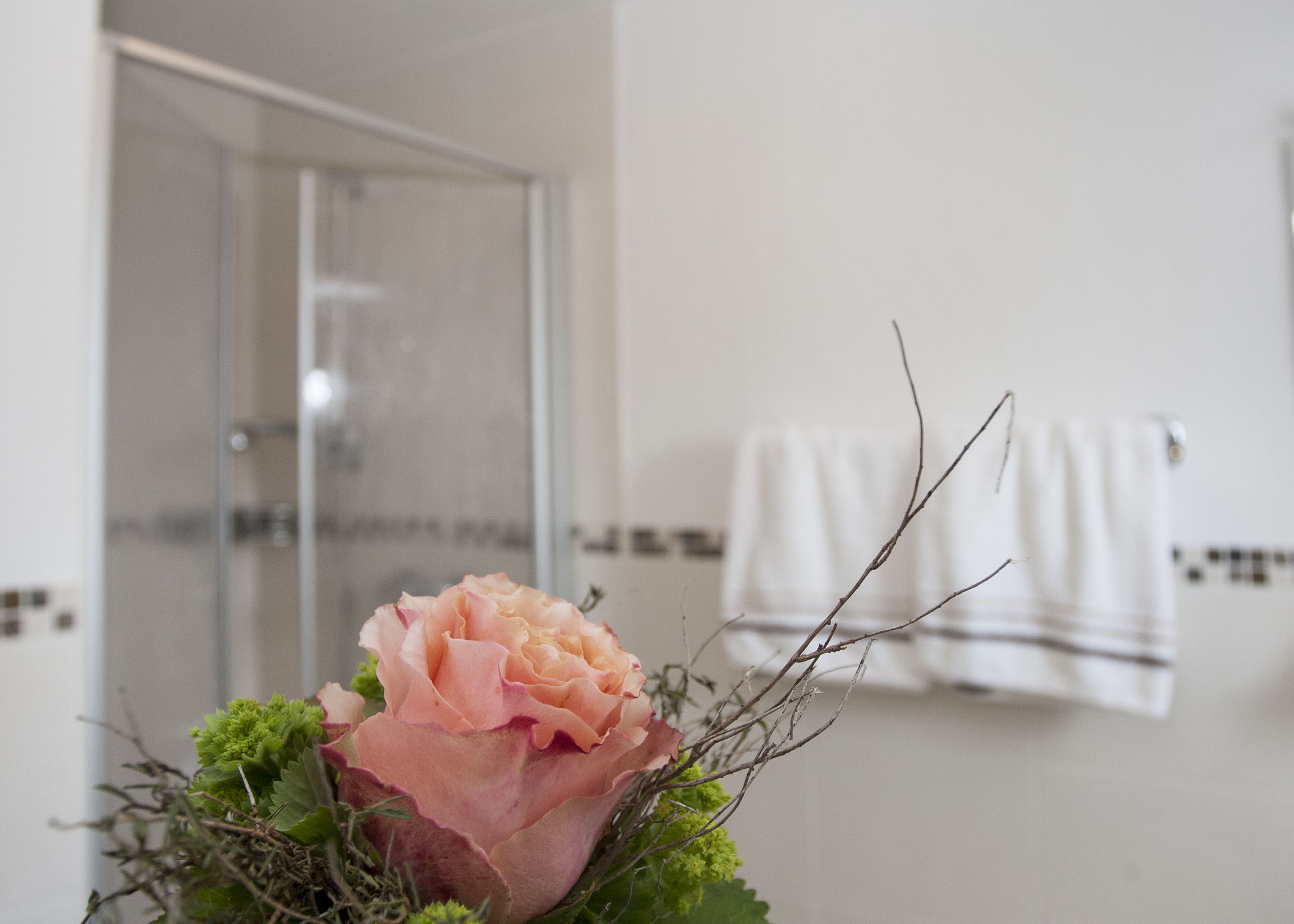 A bathroom with a glass shower door, a white towel hanging on a rack, and a pink flower in a vase.