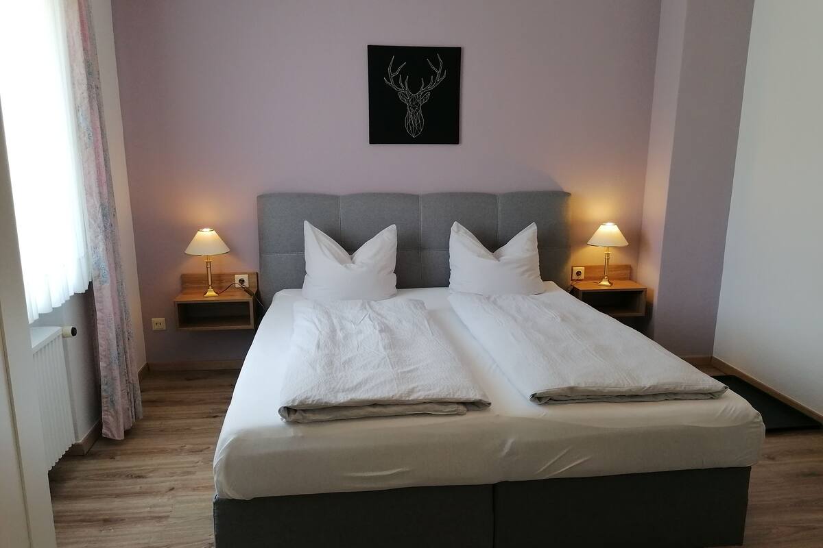 Ein ordentlich bezogenes Bett mit weißen Bettwäsche, zwei Kissen und einem Kopfteil. Auf den hölzernen Nachttischen stehen jeweils zwei Nachttischlampen. Über dem Bett hängt ein Bild mit einem Hirschkopf.
