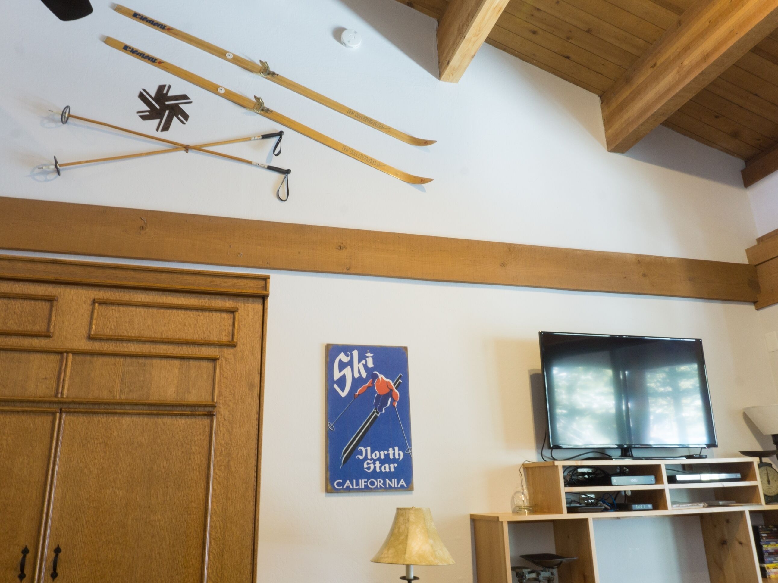 Une pièce avec des poutres en bois, un téléviseur fixé au mur, une affiche sur le thème du ski et des paires de skis accrochés au mur.